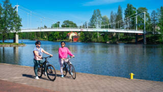 Kaksi naista pyöräilee Valkeakosken keskustassa järven rannassa.