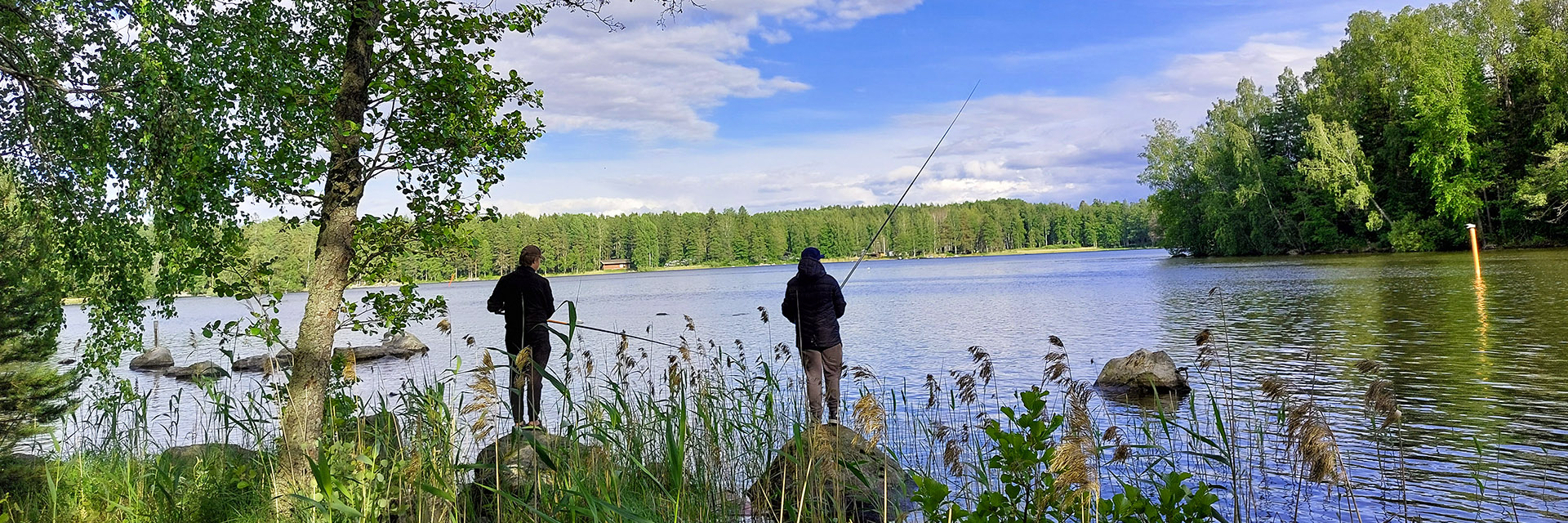 Kaksi poikaa kalastaa mato-ongilla järvenrannala.